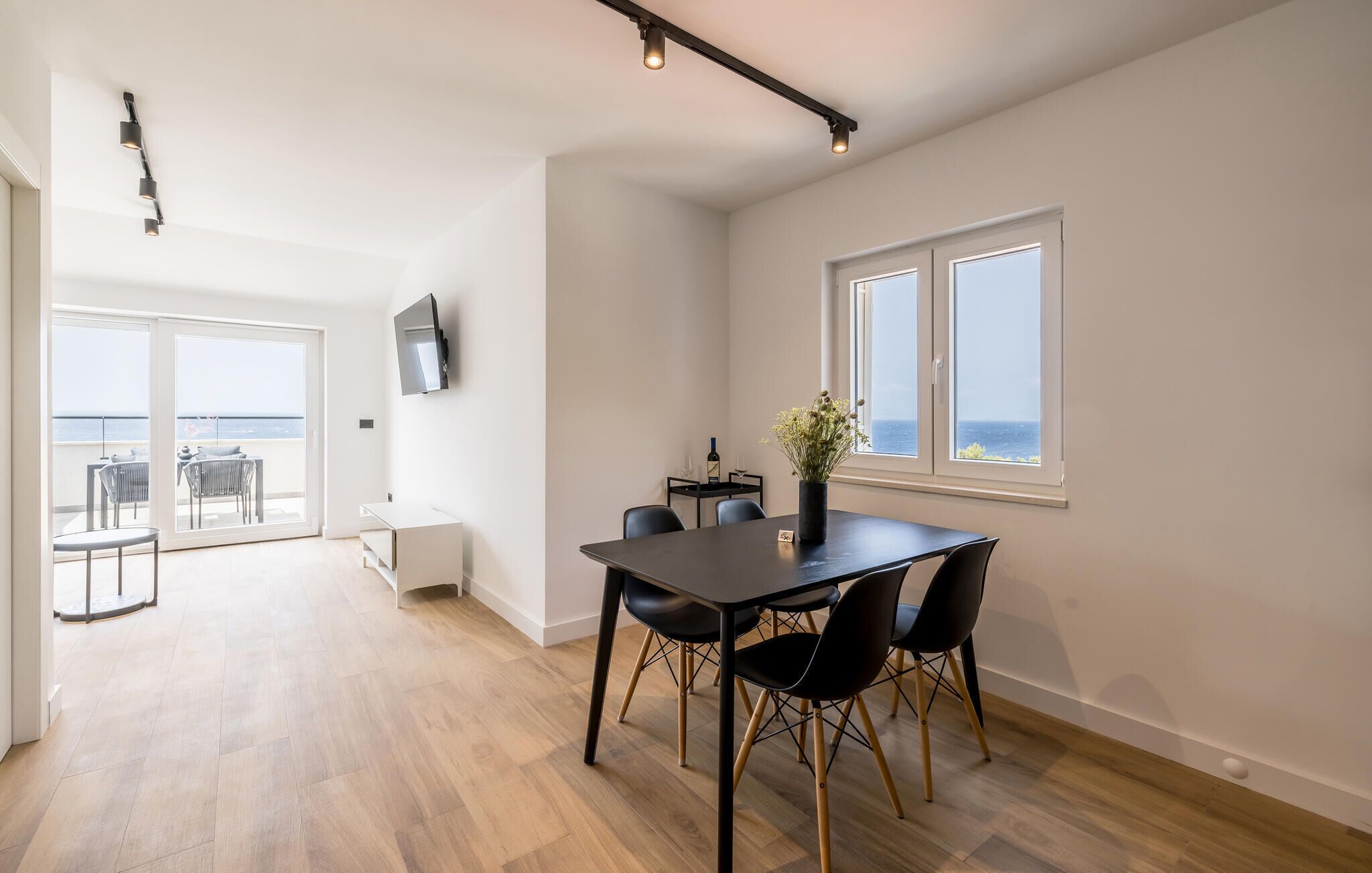 Un espace repas moderne avec une table et des chaises, un téléviseur à écran plat fixé au mur et une vue sur la mer grâce à de grandes fenêtres.