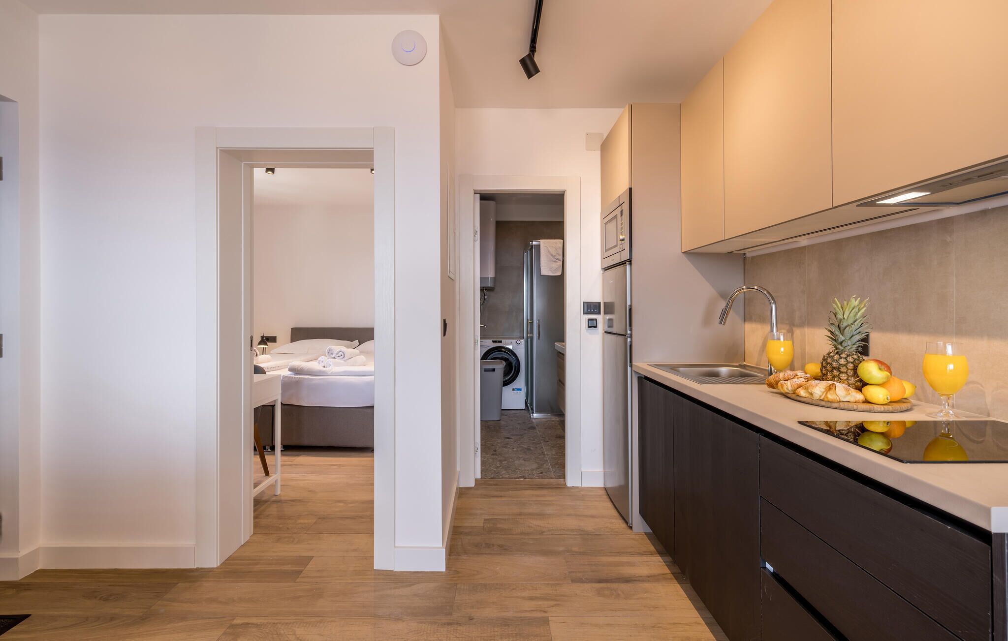 A modern hotel room with a neatly made bed, a washing machine, and a kitchen area with fresh fruits and a glass of orange juice.
