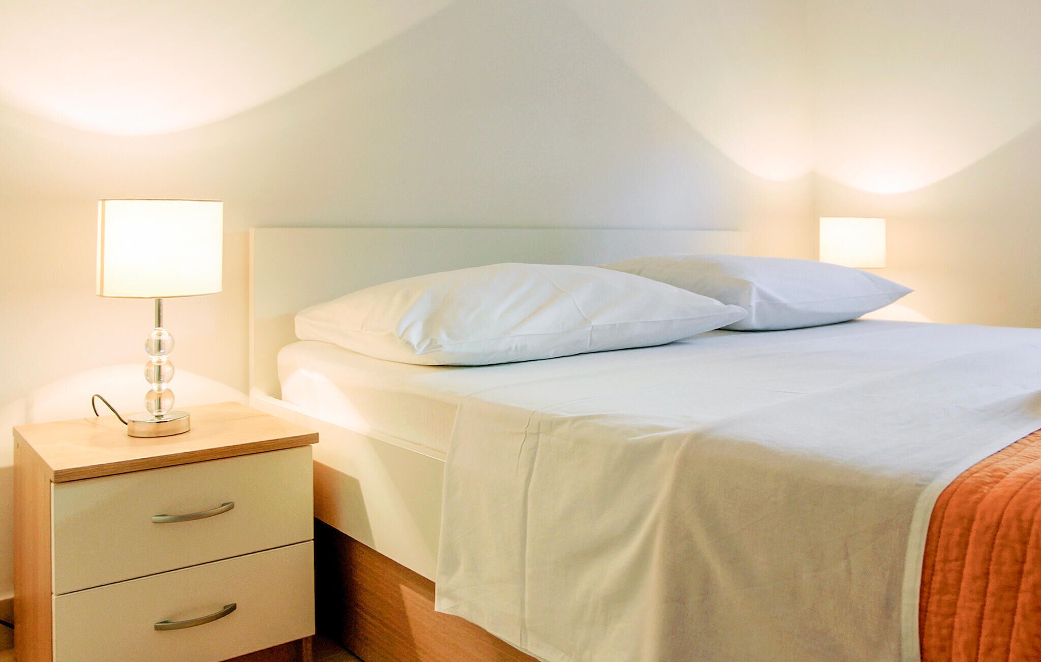 A neatly made bed with white linens and pillows, a bedside table with a lamp, and a wooden nightstand.