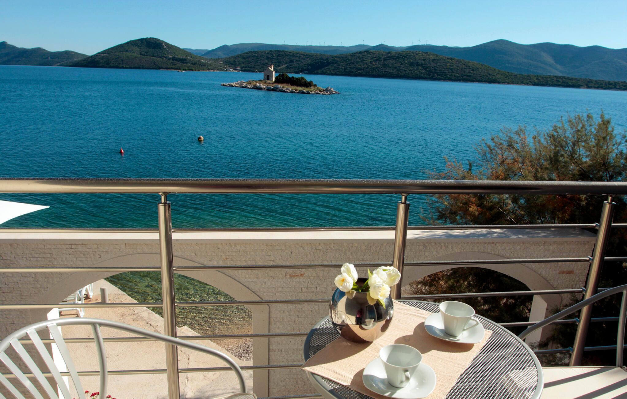 Un balcon avec une table dressée pour deux personnes, donnant sur un lac avec une petite île et des montagnes à l’horizon.