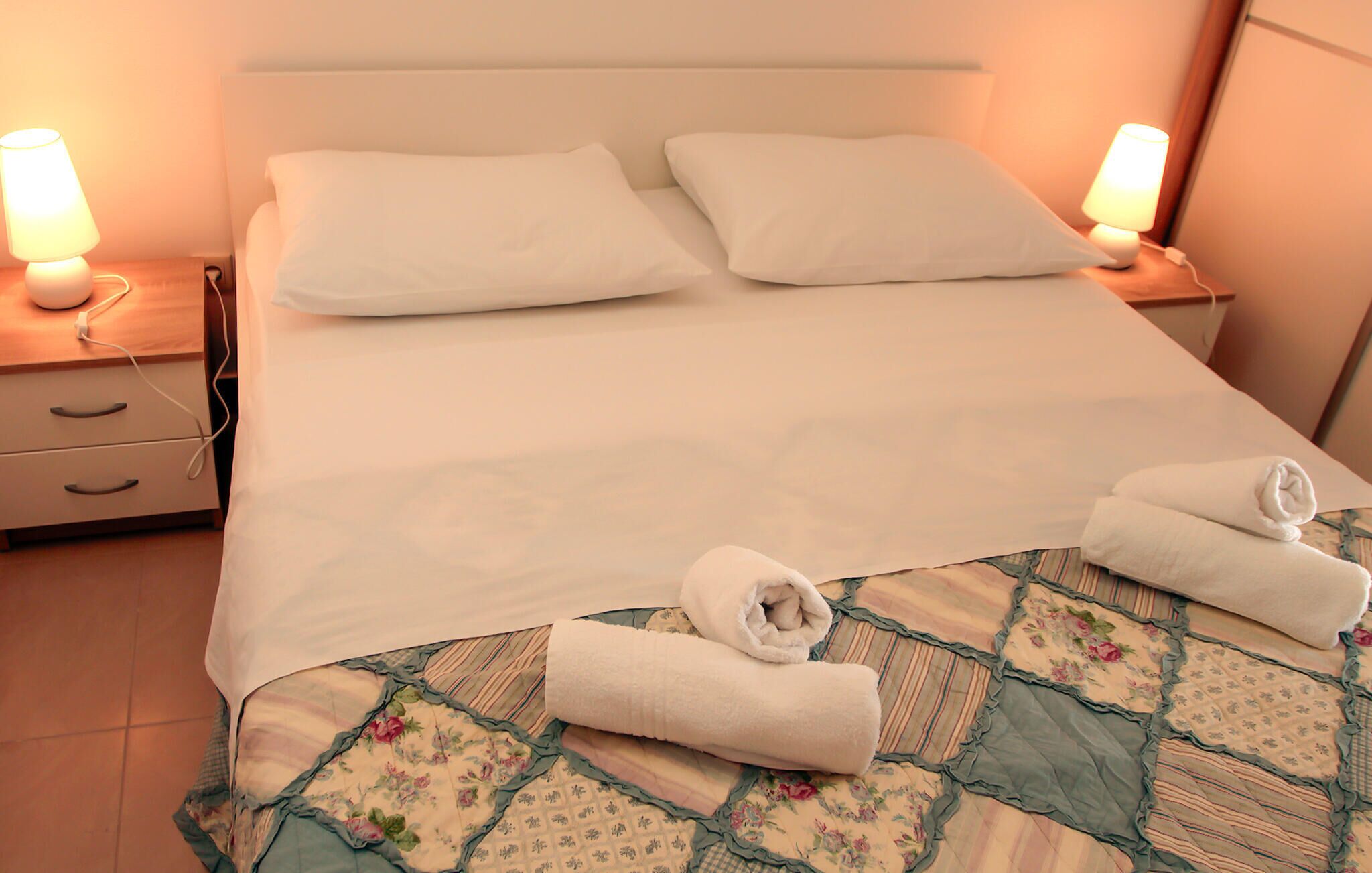 A neatly made bed with white linens, two bedside lamps, a wooden nightstand, and a floral-patterned bedspread.