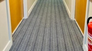 En hotellkorridor med heltäckningsmatta på golvet, trädörrar och en brandsläckare på höger sida.