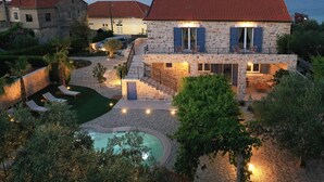 Una casa de piedra de dos pisos con techo de tejas rojas, piscina y mobiliario de exterior.