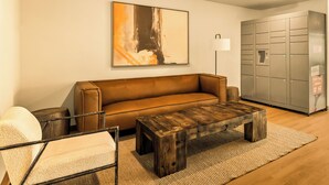 A modern living room with a brown leather sofa, a wooden coffee table, a white armchair, and a large abstract painting on the wall. There is a grey storage unit with lockers and a floor lamp. - LivAway Suites Austin San Marcos (San Marcos)