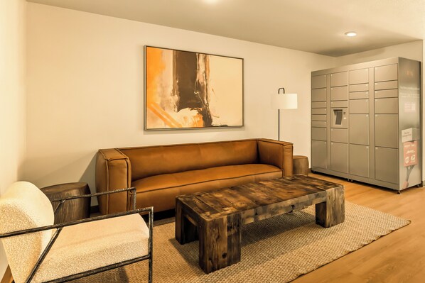 Un salon moderne avec un canapé en cuir marron, une table basse en bois, un fauteuil blanc et un grand tableau abstrait accroché au mur. On y trouve également un meuble de rangement gris avec des casiers et un lampadaire.