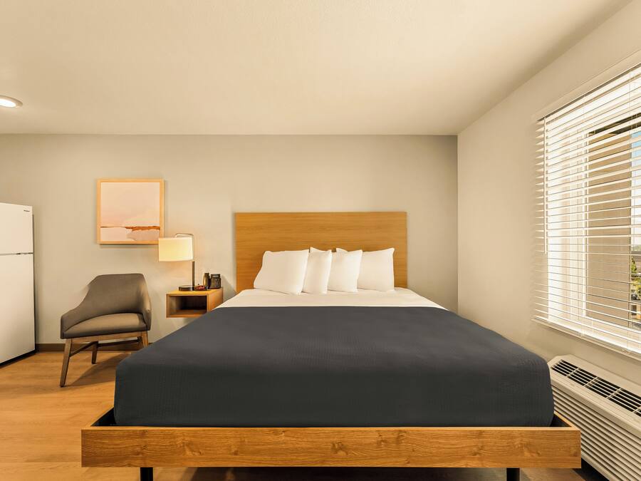 A hotel room with a bed, a chair, a desk with a lamp, and a window with blinds.