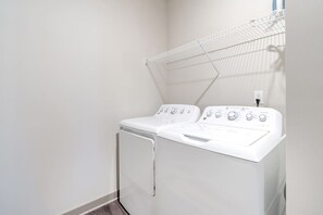 A laundry room with a front-loading washer and dryer, a drying rack, and a power outlet on the wall.