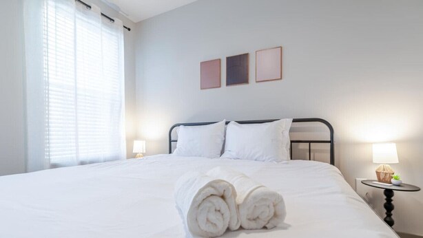 Una cama bien tendida con sábanas blancas y una toalla enrollada en el centro, una mesita de noche con una lámpara y una ventana con persianas.