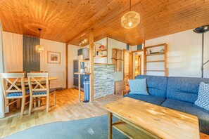 A cozy room with a wooden ceiling, a blue sofa, a wooden dining table with chairs, and a kitchen area with a stove and cabinets. - Binzer Waldhof - Haus Fuchsbau (Binz)