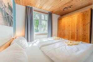 A bedroom with two beds, a window with curtains, a wooden wardrobe, and a ceiling with exposed beams. - Binzer Waldhof - Haus Fuchsbau (Binz)