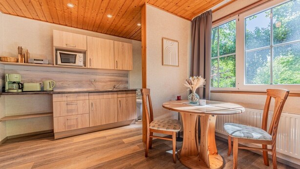A wooden-paneled kitchen with a round wooden table and chairs, a window with curtains, and a view of greenery outside. - Binzer Waldhof - Igelhaus (Binz)