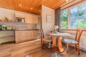 A wooden-paneled kitchen with a round wooden table and chairs, a window with curtains, and a view of greenery outside. - Binzer Waldhof - Igelhaus (Binz)