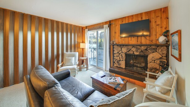 A living room with a stone fireplace, wooden panel walls, a leather sofa, and a television mounted above the fireplace. - Walk to Chair Lift: Mtn-View Schweitzer Studio! (Sandpoint)