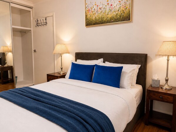 Una cama bien hecha con una manta azul, dos lámparas de noche y un cuadro de flores en la pared.