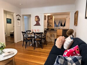 A living room with a dark sofa, a coffee table, and a dining area with a table and chairs. - A Little Home close to Beach & F1 withPrivate Courtyard Free Parking, Ground fl (St Kilda)