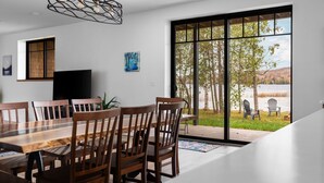 A dining area with a large table and chairs, a TV, and a view of trees and water outside. - [Modern Lake House] hot tub, dock, sandy shoreline, kayaks, foosball, Bellaire. (Bellaire)
