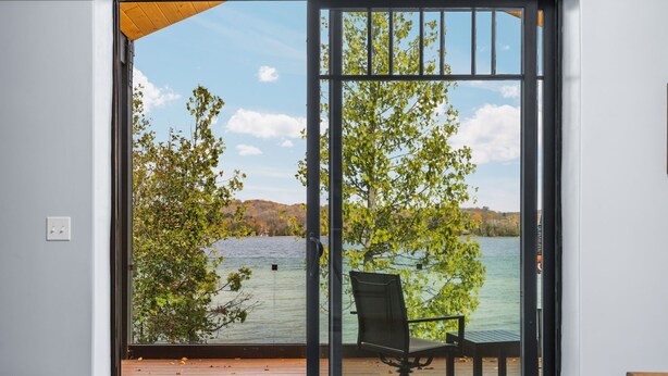 A room with a large sliding glass door leading to a wooden deck overlooking a lake with trees and hills in the background. - [Modern Lake House] hot tub, dock, sandy shoreline, kayaks, foosball, Bellaire. (Bellaire)