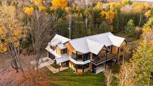 A modern two-story house with a large deck and a covered porch, surrounded by autumn foliage. - [Modern Lake House] hot tub, dock, sandy shoreline, kayaks, foosball, Bellaire. (Bellaire)