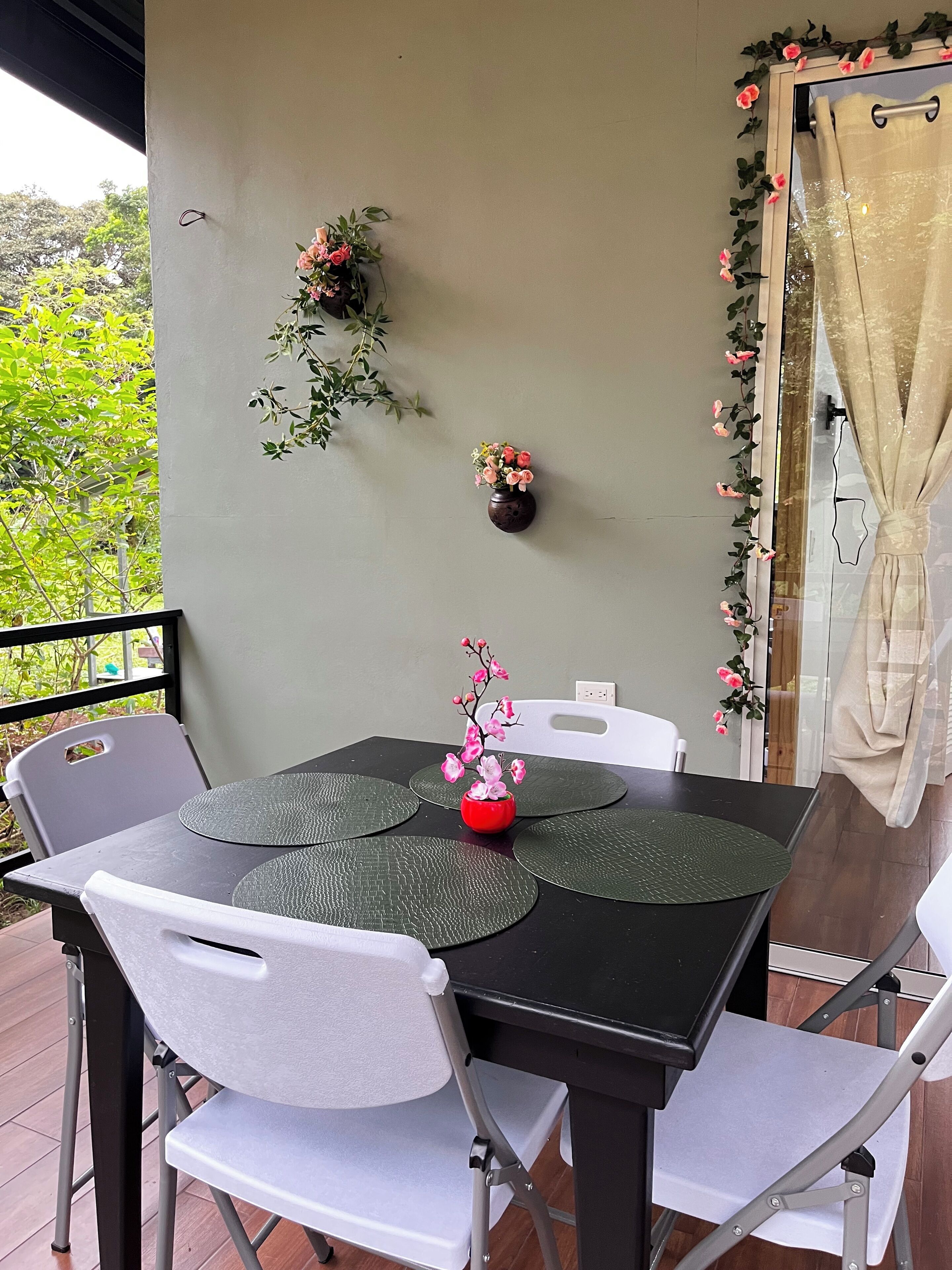 Un comedor con una mesa negra, sillas blancas y un florero rojo con flores rosadas.