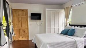 A bedroom with a wooden door, a bed with white bedding and blue pillows, a mounted TV, and a white air conditioning unit. - Toucan House (Valle Azul)