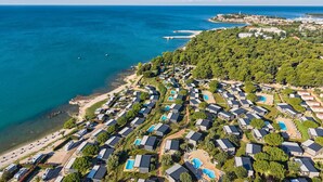 Eine Luftaufnahme eines Küstenresorts mit mehreren bungalowsähnlichen Unterkünften, einem zentralen Swimmingpool sowie Blick auf das Meer und eine in der Ferne liegende Stadt.