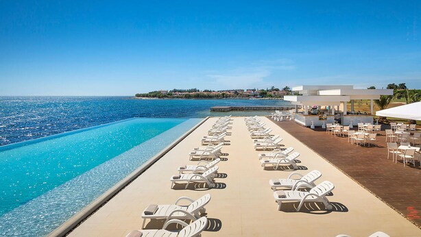 Ein Poolbereich mit Liegestühlen und einer Bar mit Blick auf das klare, blaue Meer.
