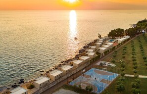 Eine Küstenlandschaft mit einer Reihung von Strandbungalows, einem Basketballplatz und Blick auf das Meer bei Sonnenuntergang.