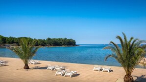 Blick auf einen Strand mit blauem Himmel, ruhiger See und einer Reihe weißer Liegestühle am Ufer.