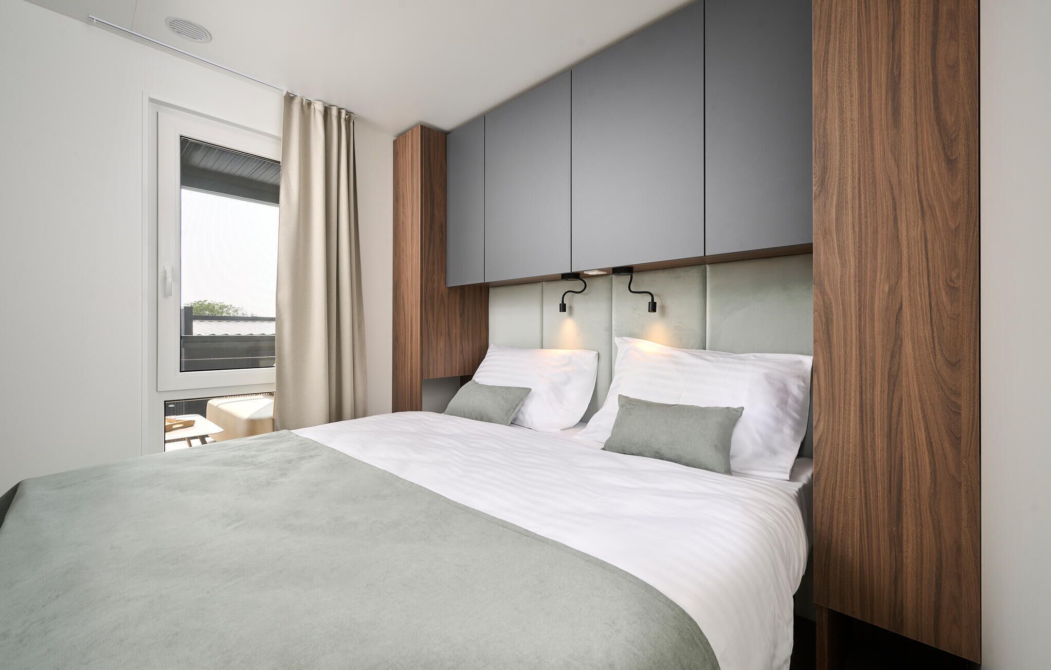 Camera da letto moderna con un letto grande, biancheria grigio e bianca, pannelli di legno e vista dalla finestra.