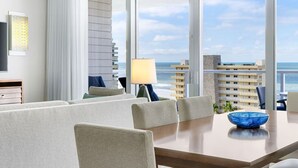 A modern hotel room with a large window offering a view of the ocean and buildings. There is a dining area with a table and chairs, a lamp, and a TV on the wall. - 12/21-26 Marriott Luxury 2-BR Beach Villa - Marco Island Resort - Crystal Shores (Marco Island)