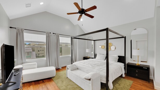 A modern bedroom with a four-poster bed, a ceiling fan, a TV, and a large window with curtains. - The Wags | Sleeps 10 (Houston)