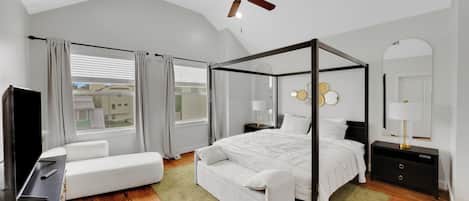 A modern bedroom with a four-poster bed, a ceiling fan, a TV, and a large window with curtains.
