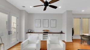 A modern living room with a ceiling fan, white sofas, a wooden dining table, and a kitchen area with white cabinets and a wooden countertop. - The Wags | Sleeps 10 (Houston)