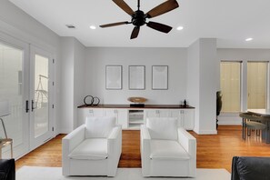 Ein modernes Wohnzimmer mit Deckenventilator, weißen Sofas, einem Holzessttisch und einer Küchenzeile mit weißen Schränken und einer Holz Arbeitsplatte.