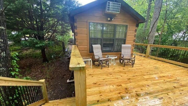A wooden deck with two chairs and a small table, leading to a cabin with a large window and a fireplace.