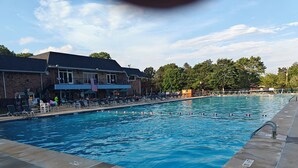 A swimming pool with lounge chairs and umbrellas, a brick building with an American flag, and trees in the background.