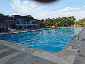 A swimming pool with lounge chairs and umbrellas, a brick building with an American flag, and trees in the background.