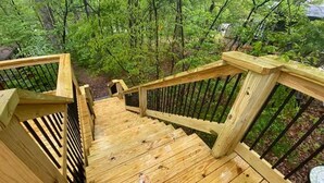 A wooden outdoor staircase leading to a deck with a metal railing, surrounded by lush greenery.