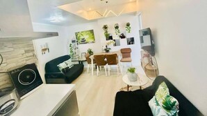 Una sala de estar moderna con un sofá negro, una mesa de comedor con sillas y un televisor. Hay plantas en las paredes y un aplique de techo.