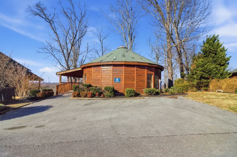 A wooden cabin with a green roof, surrounded by trees and a paved driveway.