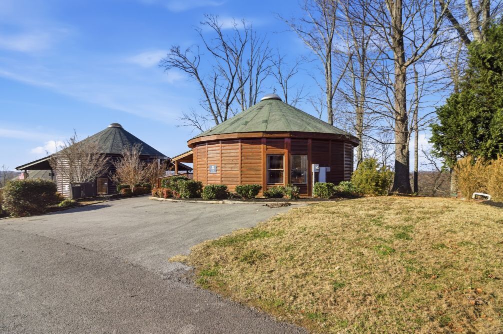 A wooden yurt with a green roof, surrounded by trees and a paved driveway.