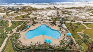 Vista aérea de um resort com uma piscina central, rodeada por palmeiras e uma praia.