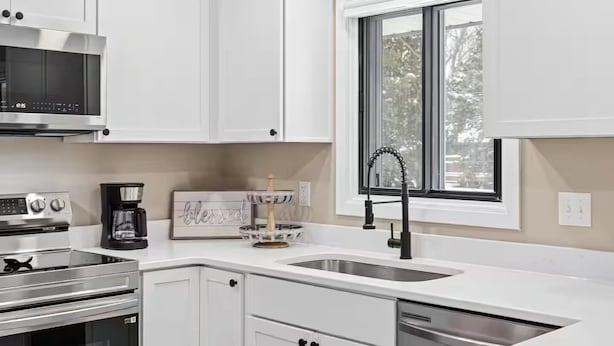 Una cocina moderna con armarios blancos, electrodomésticos de acero inoxidable, un lavamanos doble y un ventanal con vista a árboles.