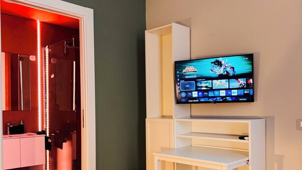 A modern room with a TV mounted on a white entertainment unit, a ceiling-mounted air conditioner, and a visible bathroom with red lighting. - Le dimore di corso Torino  (Novara)