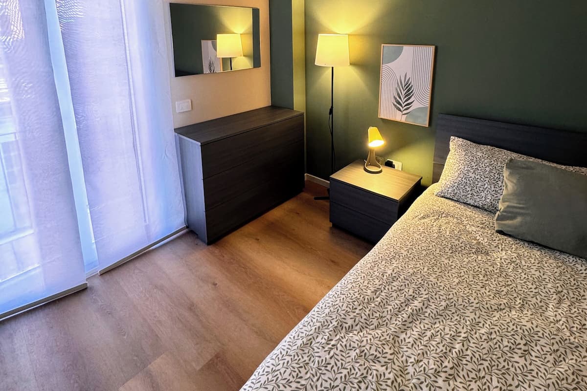 A hotel room with a bed, a bedside table with a lamp, a mirror, and a window with sheer curtains.