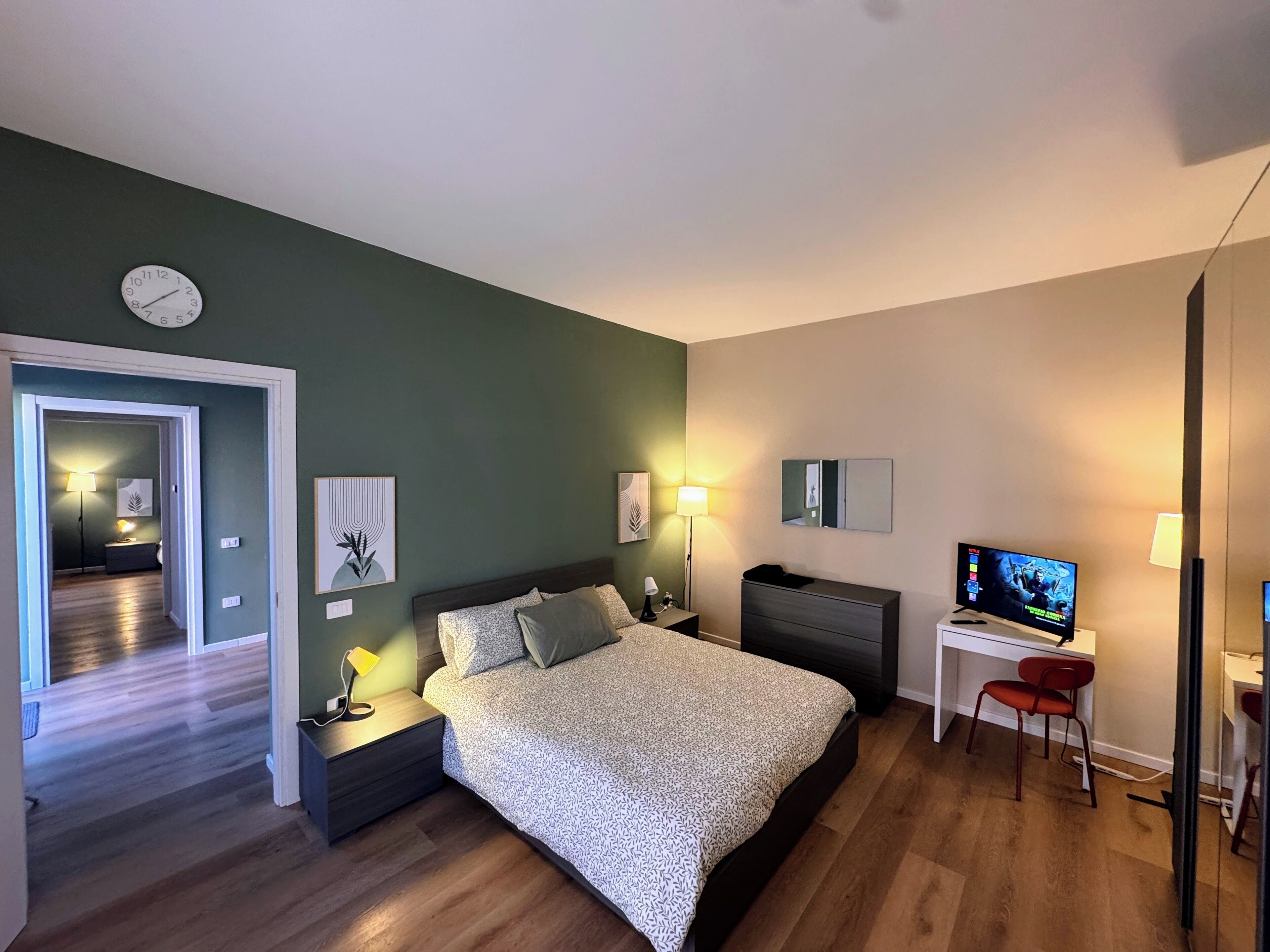 Ein modernes Schlafzimmer mit Bett, Nachttischen, Schreibtisch, Stuhl, Fernseher und einer Wanduhr.
