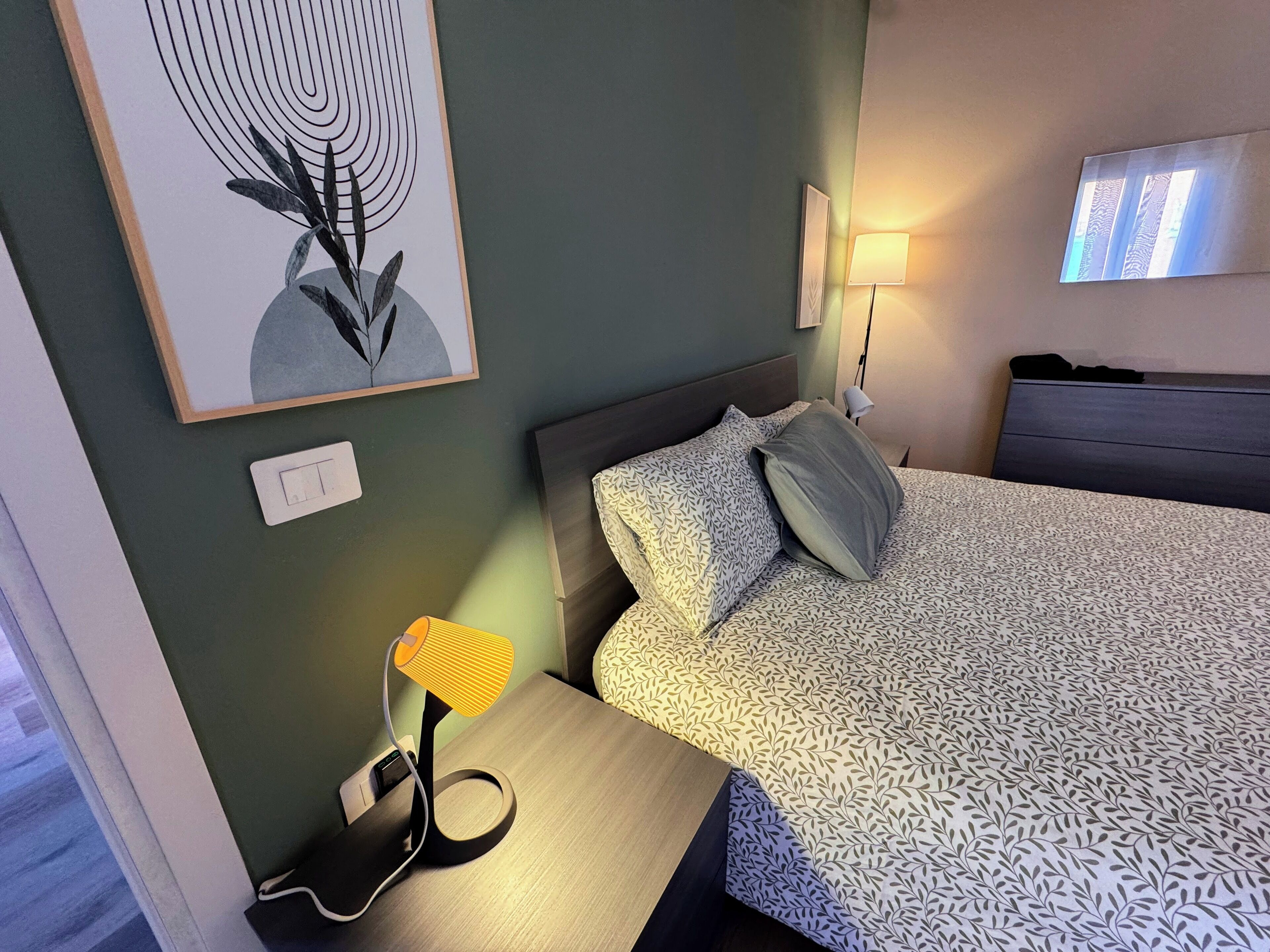 Habitación de hotel con cama, mesita de noche con lámpara, luz empotrada en la pared y una obra de arte enmarco.