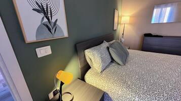 Habitación de hotel con cama, mesita de noche con lámpara, luz empotrada en la pared y una obra de arte enmarco.