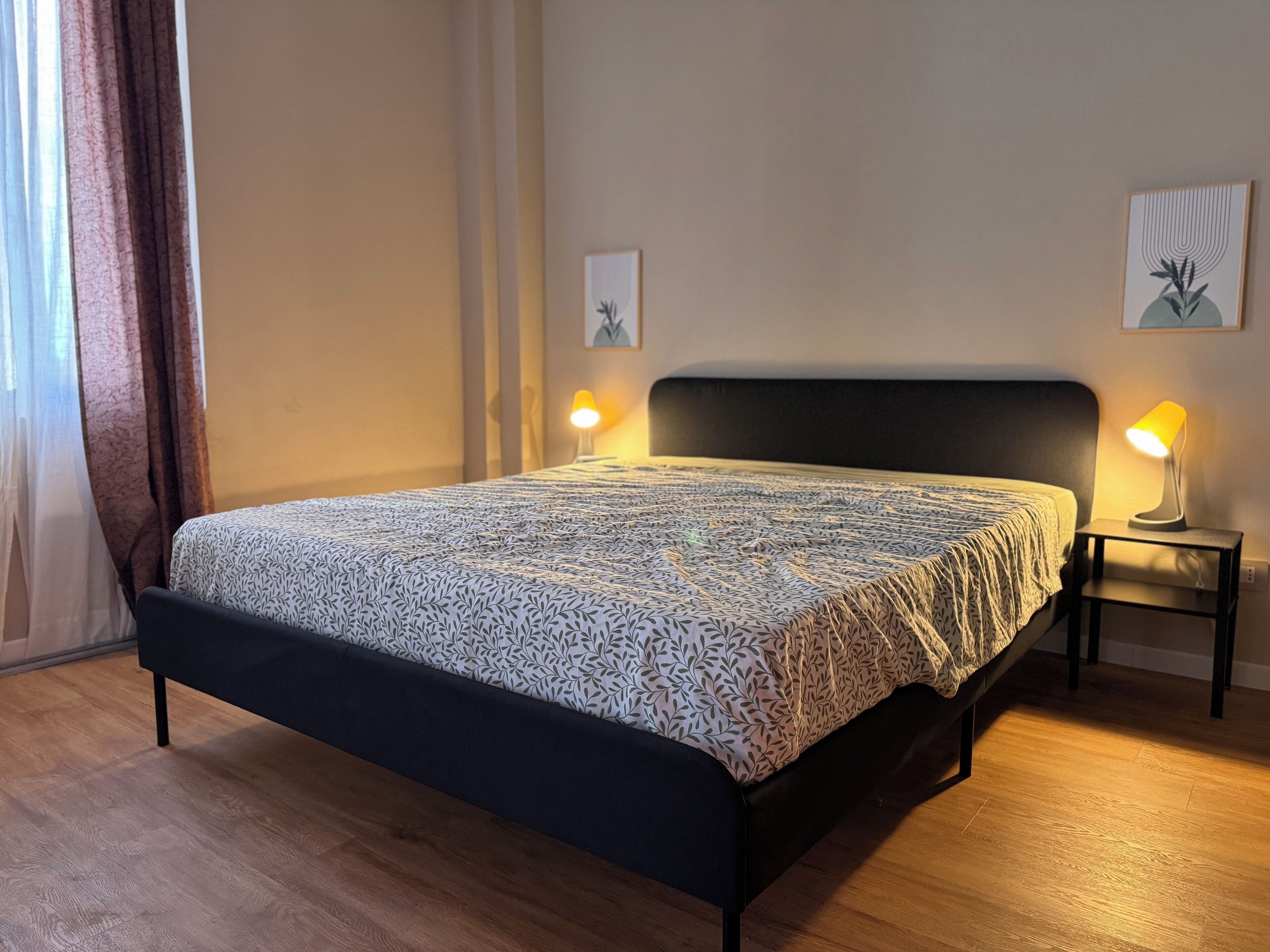 Une chambre à coucher comprenant un lit, deux lampes de chevet, une fenêtre avec des rideaux et deux œuvres d’art accrochées au mur.