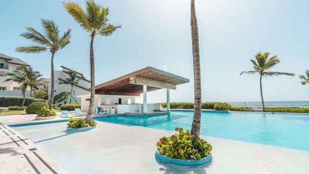 Ein Poolbereich mit Palmen, einer überdachten Lounge und Blick aufs Meer.